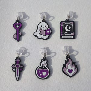 Pode incluir: Um conjunto de seis pingentes com uma chave de coração, um fantasma lendo um livro, um livro com lua crescente, uma espada com coração, um frasco de poção e uma chama em forma de coração. Cada pingente é nas cores roxo, branco e preto.
