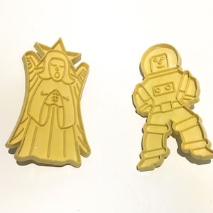 Peut inclure: Deux emporte-pièces jaunes : l'un en forme d'ange avec une couronne en étoile et des ailes déployées, et l'autre un astronaute rétro en combinaison spatiale. Les deux sont faits d'un matériau solide de couleur claire, sur fond blanc.