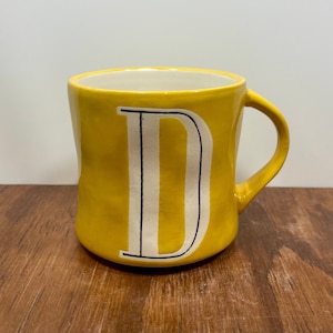 May include: A yellow ceramic mug with a white interior and a large white letter "D" with black outlines. The mug has a rounded shape and a curved handle. The mug is sitting on a wooden surface.