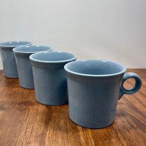 May include: A set of four light blue ceramic mugs arranged in a diagonal line. Each mug features a smooth, solid-coloured finish, a cylindrical body with a slightly flared rim, and a small, circular handle attached to the side of the vessel.