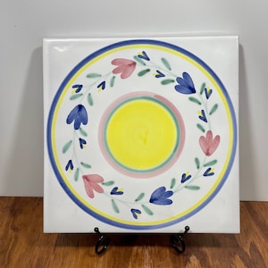 May include: Square white ceramic tile with a hand-painted floral design. The tile features a central yellow circle, surrounded by pink and green rings, and a blue and yellow border. Pink and blue flowers are arranged around the central circle.