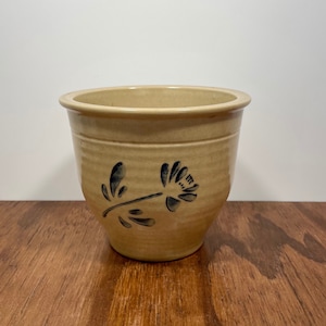 May include: A tan ceramic planter with a floral design. The planter has a rounded shape with a slightly flared rim. A dark blue floral pattern is painted on the side. The planter sits on a wooden surface.