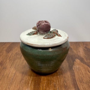 May include: A handcrafted ceramic jar with a lid. The jar is a deep green color, with a white lid and a purple rose-shaped handle. The lid has green and brown leaf accents. The jar is sitting on a wooden surface.