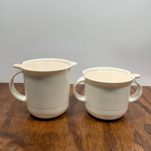 May include: Two off-white, lidded serving pitchers with handles. The larger pitcher has a spout. The lids are a light tan color. The pitchers are on a dark wood surface.