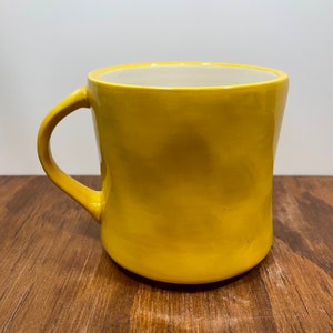 Peut inclure: Une tasse en c&eacute;ramique jaune avec une anse incurv&eacute;e. La tasse a une finition lisse et brillante et un int&eacute;rieur de couleur cr&egrave;me. La tasse est pos&eacute;e sur une surface en bois.