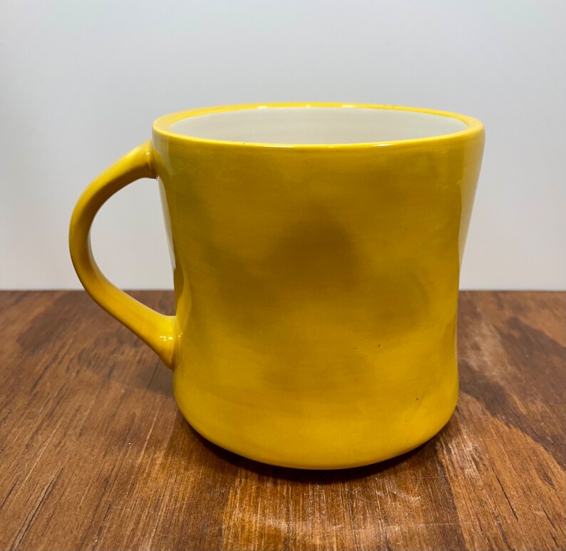 Peut inclure: Une tasse en c&eacute;ramique jaune avec une anse incurv&eacute;e. La tasse a une finition lisse et brillante et un int&eacute;rieur de couleur cr&egrave;me. La tasse est pos&eacute;e sur une surface en bois.