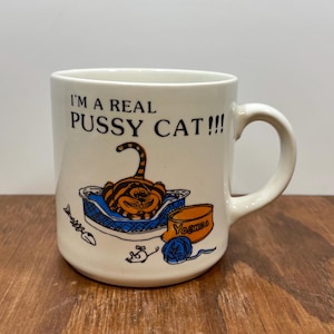 May include: Cream-colored ceramic mug with the text "I'M A REAL PUSSY CAT!!!" and a cartoon cat in a blue bed. The mug also features a cat food bowl, a ball of yarn, and a fishbone design. The mug has a curved handle.