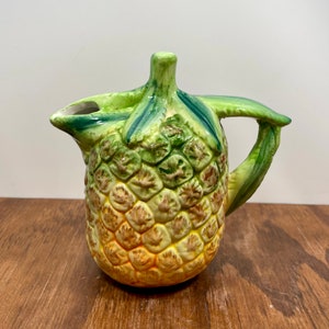 May include: A ceramic pineapple-shaped pitcher with a green handle and spout. The pitcher's body is yellow and brown, with green leaves at the top. The pitcher is designed to resemble a pineapple.