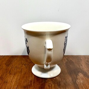 Puede incluir: Taza de t&eacute; color crema con base de pedestal y asa curva. La taza presenta patrones decorativos negros alrededor del borde. La taza est&aacute; sobre una superficie de madera.