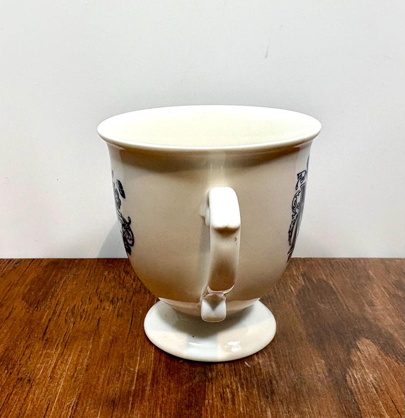 Puede incluir: Taza de t&eacute; color crema con base de pedestal y asa curva. La taza presenta patrones decorativos negros alrededor del borde. La taza est&aacute; sobre una superficie de madera.