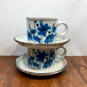 May include: Two stacked ceramic coffee cups with matching saucers. The cups and saucers are off-white with a speckled texture and a dark brown rim. Each cup is decorated with blue floral designs.