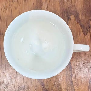 Puede incluir: Vista a&eacute;rea de una taza de t&eacute; de cer&aacute;mica blanca con asa. La taza est&aacute; vac&iacute;a y se encuentra sobre una superficie de madera. El interior de la taza es liso y el asa es curva.