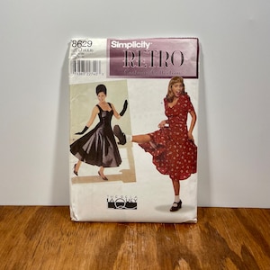 Puede incluir: Un patrón de costura Simplicity Retro Costume Collection. El paquete presenta imágenes de dos vestidos vintage. La parte superior del paquete tiene el número 8629 y el logotipo de Simplicity. El paquete está sobre una superficie de madera.