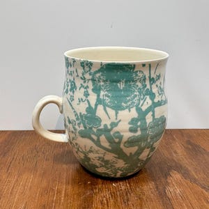 K&ouml;nnte beinhalten: Eine Keramik-Tasse mit cremefarbenem Grund und einem t&uuml;rkisfarbenen Blumenmuster. Die Tasse hat einen abgerundeten K&ouml;rper, einen gebogenen Henkel und eine weite &Ouml;ffnung. Das florale Design umfasst Zweige und Bl&auml;tter.