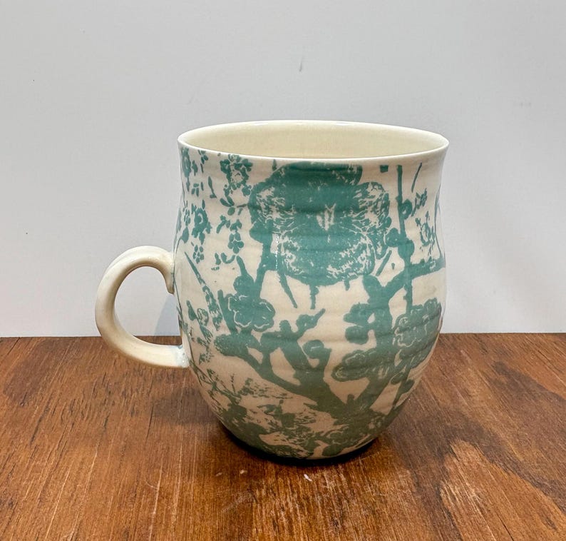 K&ouml;nnte beinhalten: Eine Keramik-Tasse mit cremefarbenem Grund und einem t&uuml;rkisfarbenen Blumenmuster. Die Tasse hat einen abgerundeten K&ouml;rper, einen gebogenen Henkel und eine weite &Ouml;ffnung. Das florale Design umfasst Zweige und Bl&auml;tter.
