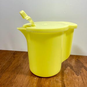 May include: A bright yellow plastic pitcher with a matching lid and handle. The pitcher has a curved design and a spout for pouring. The lid is open, revealing the interior. The pitcher is sitting on a wooden surface.