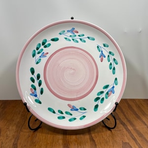 May include: A white ceramic plate with a pink rim and center, adorned with green leaves and blue and pink flowers. The plate is displayed on a black metal stand, showcasing its decorative design.