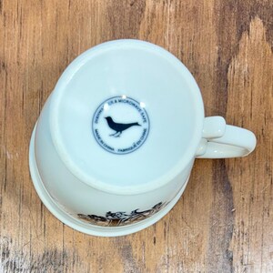 Puede incluir: Taza de t&eacute; de cer&aacute;mica blanca con asa, con un dise&ntilde;o decorativo en el borde inferior. La parte inferior de la taza tiene un emblema circular con una silueta de p&aacute;jaro y el texto "Dishwasher & Microwave Safe, Made in China, Fabriqu&eacute; en Chine."