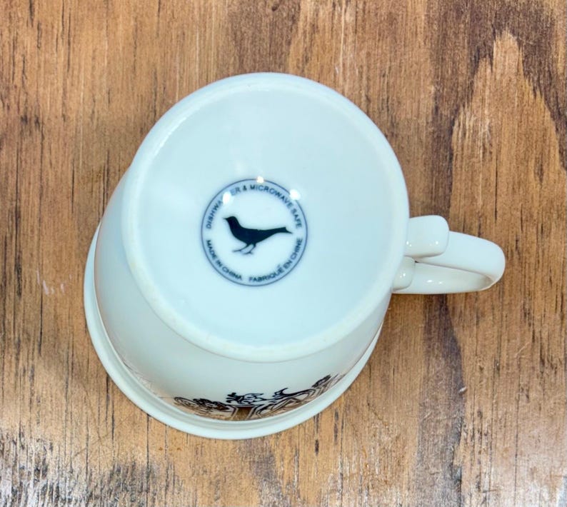 Puede incluir: Taza de t&eacute; de cer&aacute;mica blanca con asa, con un dise&ntilde;o decorativo en el borde inferior. La parte inferior de la taza tiene un emblema circular con una silueta de p&aacute;jaro y el texto "Dishwasher & Microwave Safe, Made in China, Fabriqu&eacute; en Chine."
