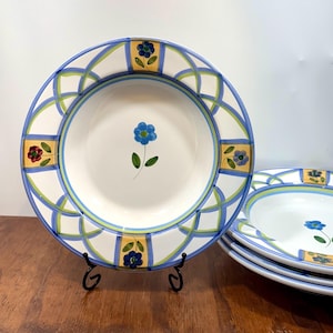 Può includere: Un set di ciotole in ceramica bianca con un motivo floreale blu al centro. Il bordo è decorato con un motivo ripetitivo di linee blu e verdi intrecciate, intervallate da pannelli rettangolari gialli contenenti piccoli disegni floreali.