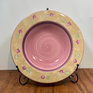 Könnte beinhalten: Ein dekorativer Keramikteller mit rosa Mitte und beigem Rand. Der Rand ist mit bemalten Radieschen und weißen Punkten verziert. Der Teller wird auf einem schwarzen Metallständer präsentiert, der sein Design hervorhebt.