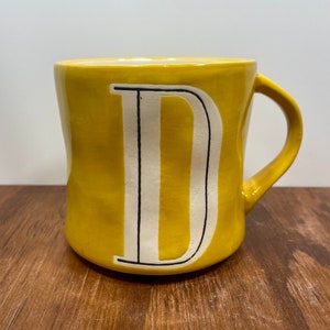Peut inclure: Une tasse en c&eacute;ramique jaune avec un grand "D" blanc au centre, soulign&eacute; de noir. La tasse a une anse incurv&eacute;e et repose sur une surface en bois. Id&eacute;ale pour le caf&eacute; ou le th&eacute;.