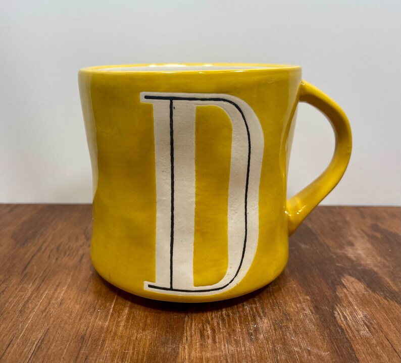Peut inclure: Une tasse en c&eacute;ramique jaune avec un grand "D" blanc au centre, soulign&eacute; de noir. La tasse a une anse incurv&eacute;e et repose sur une surface en bois. Id&eacute;ale pour le caf&eacute; ou le th&eacute;.