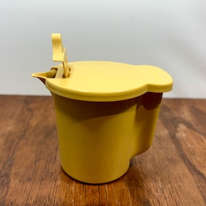May include: A yellow plastic container with a lid and a handle. The container has a rounded shape and a spout. The lid is attached to the container with a hinge. The handle is raised. The container is sitting on a wooden surface.