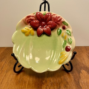 May include: A small, scalloped ceramic dish with a pale green center and a raised, three-dimensional dark red flower at the top. The rim is decorated with embossed green leaves, yellow accents, and small red berries on a cream-colored background.
