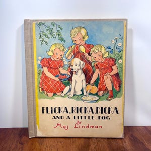 May include: A hardback book titled Flicka, Ricka, Dicka and a Little Dog by Maj Lindman. The cover features an illustration of three blonde children in red plaid dresses interacting with a white and brown dog in a grassy field with flowers.