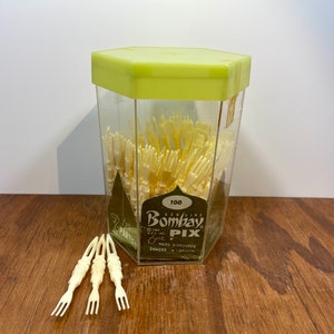 Peut inclure: Un récipient vintage de fourchettes à cocktail "Bombay Pix". Le récipient en plastique transparent a un couvercle hexagonal vert clair et contient de nombreuses fourchettes en plastique blanc cassé. L'étiquette indique "100" et "Hors d'oeuvres Snacks Drinks". Trois fourchettes sont à l'extérieur.