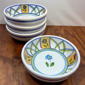 Può includere: Un set di quattro ciotole in ceramica bianca con un motivo floreale dipinto a mano. Ogni ciotola presenta un fiore blu centrale, circondato da pannelli gialli con foglie verdi e bordi a griglia blu. I bordi esterni sono rifiniti con fasce blu.