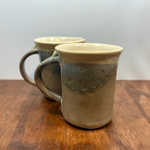 Puede incluir: Dos tazas de cerámica hechas a mano con un esmalte moteado. Las tazas tienen un borde beige, una banda azul-verdosa y una base marrón. Cada taza tiene un asa curva. Las tazas están sobre una superficie de madera.