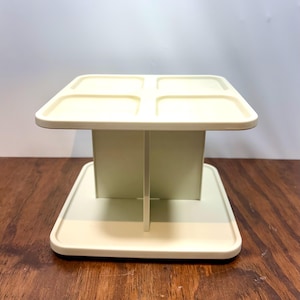 May include: A cream-colored plastic organizer stand featuring a square base and a top platform divided into four distinct square compartments. The structure is supported by a central vertical divider that connects the base to the top tier.