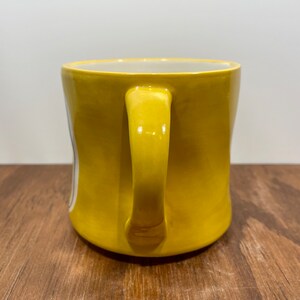 Peut inclure: Une tasse en c&eacute;ramique jaune avec une anse incurv&eacute;e. La tasse a une finition brillante et un int&eacute;rieur blanc. La tasse est pos&eacute;e sur une surface en bois.