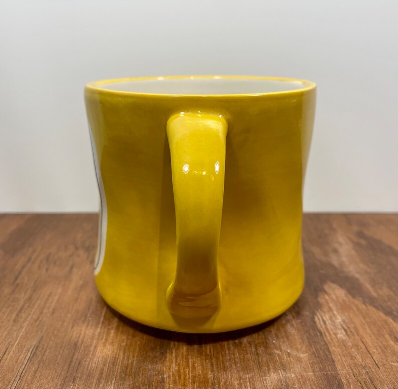 Peut inclure: Une tasse en c&eacute;ramique jaune avec une anse incurv&eacute;e. La tasse a une finition brillante et un int&eacute;rieur blanc. La tasse est pos&eacute;e sur une surface en bois.