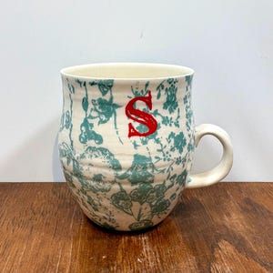 K&ouml;nnte beinhalten: Eine Keramik-Tasse mit cremefarbenem Grund und einem teal-farbenen Blumenmuster. Die Tasse hat einen roten Buchstaben "S" und einen gebogenen Henkel. Die Tasse steht auf einer Holzoberfl&auml;che.