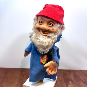 Peut inclure: Une poupée gnome vintage avec un visage souriant, un chapeau en feutre rouge et une longue barbe blanche. La poupée porte une tunique en feutre bleu avec l'étiquette "Gucki". La poupée est sur un socle blanc.