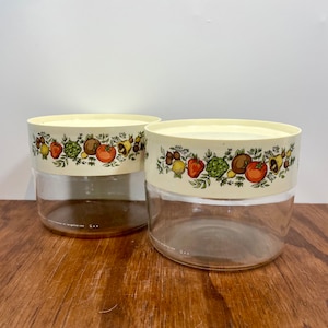 May include: Two clear glass jars with off-white lids. The lids feature a colorful border of various fruits and vegetables. The jars are sitting on a wooden surface.