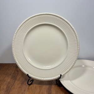 May include: A round, off-white ceramic dinner plate featuring a smooth, recessed center and a wide, raised rim decorated with vertical fluted lines and a delicate, textured wheat-like pattern along the outer edge, displayed on a black stand.