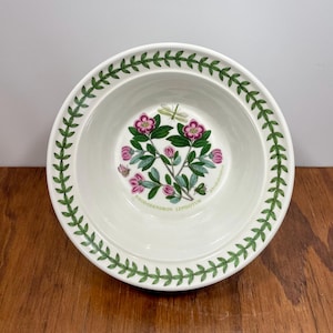 Puede incluir: Un cuenco de cerámica blanco con un borde de hojas verdes y un diseño floral en el centro. El diseño floral presenta flores rosas, hojas verdes y el texto "RHODODENDRON LEPIDOTUM".