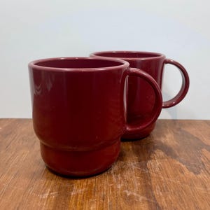 May include: Two matching deep red mugs with a smooth, glossy finish. Each mug features a cylindrical body with a slightly wider base and a curved handle on the side. The mugs are positioned one behind the other, showcasing their uniform color.