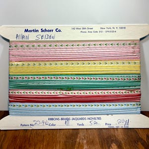 Puede incluir: Un carrete de cintas Allan Seiden de Martin Schorr Co. presenta varios colores, incluyendo rosa, amarillo, verde, rojo y azul. Las cintas tienen patrones decorativos y están etiquetadas con detalles como "RIBBONS-BRAIDS-JAQUARDS-NOVELTIES" y "20 yds."