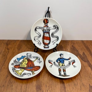 May include: Three white decorative plates with colorful illustrations. One plate features a barber pole design, another depicts a fireman, and the third shows a jockey. Each plate has text, including "Barber Pole," "Fireman," and "Jockey Hitching Post."