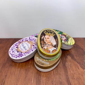 Puede incluir: Cuatro latas ovaladas de pastillas Flavigny. Una lata es blanca con diseños florales morados y la palabra "Violet". Otra lata es dorada con un retrato de una mujer y las palabras "La Reine Mint Pastilles". Las otras dos latas tienen diseños coloridos.