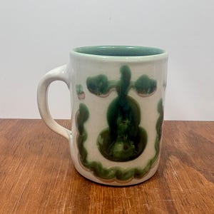 Puede incluir: Una taza de cerámica blanca con interior verde y asa. La taza presenta un diseño floral verde oscuro con un contorno verde claro. La taza está sobre una superficie de madera.