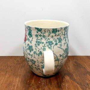 K&ouml;nnte beinhalten: Eine Keramik-Tasse mit cremefarbenem Innenraum und wei&szlig;em Henkel. Das &Auml;u&szlig;ere zeigt ein florales Muster in T&uuml;rkis und Rot auf cremefarbenem Hintergrund. Die Tasse steht auf einer Holzoberfl&auml;che.