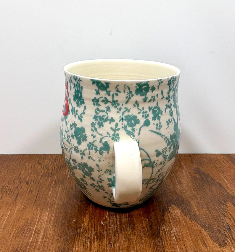 K&ouml;nnte beinhalten: Eine Keramik-Tasse mit cremefarbenem Innenraum und wei&szlig;em Henkel. Das &Auml;u&szlig;ere zeigt ein florales Muster in T&uuml;rkis und Rot auf cremefarbenem Hintergrund. Die Tasse steht auf einer Holzoberfl&auml;che.