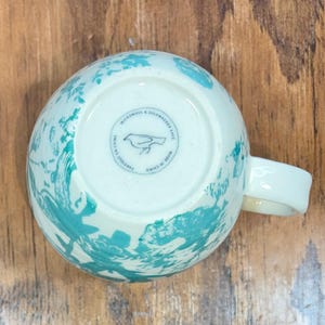 K&ouml;nnte beinhalten: Wei&szlig;e Keramik-Tasse mit einem t&uuml;rkisfarbenen, abstrakten Design. Die Tasse hat einen gebogenen Henkel und einen runden Stempel auf der Unterseite mit einer Vogelillustration und dem Text "MICROWAVE & DISHWASHER SAFE" und "MADE IN CHINA".
