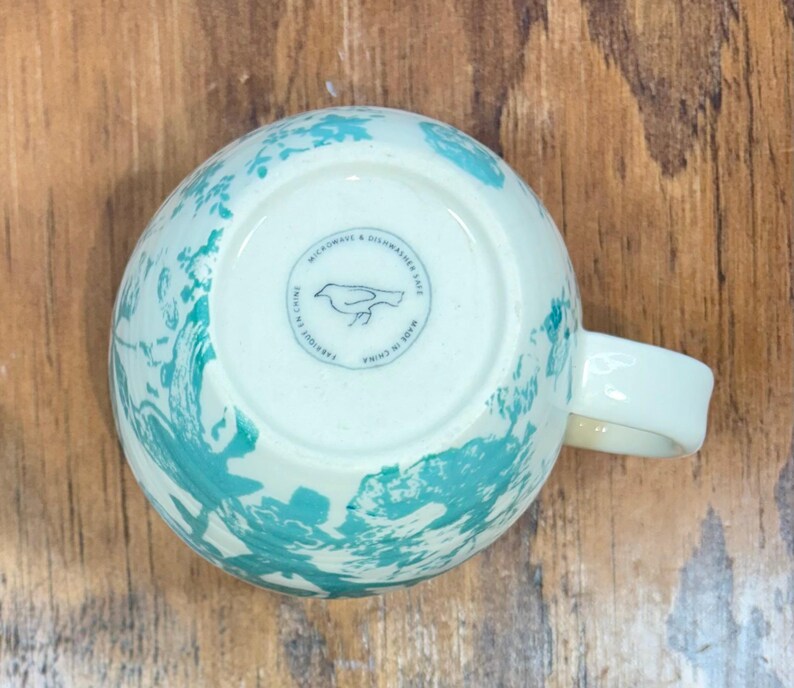 K&ouml;nnte beinhalten: Wei&szlig;e Keramik-Tasse mit einem t&uuml;rkisfarbenen, abstrakten Design. Die Tasse hat einen gebogenen Henkel und einen runden Stempel auf der Unterseite mit einer Vogelillustration und dem Text "MICROWAVE & DISHWASHER SAFE" und "MADE IN CHINA".